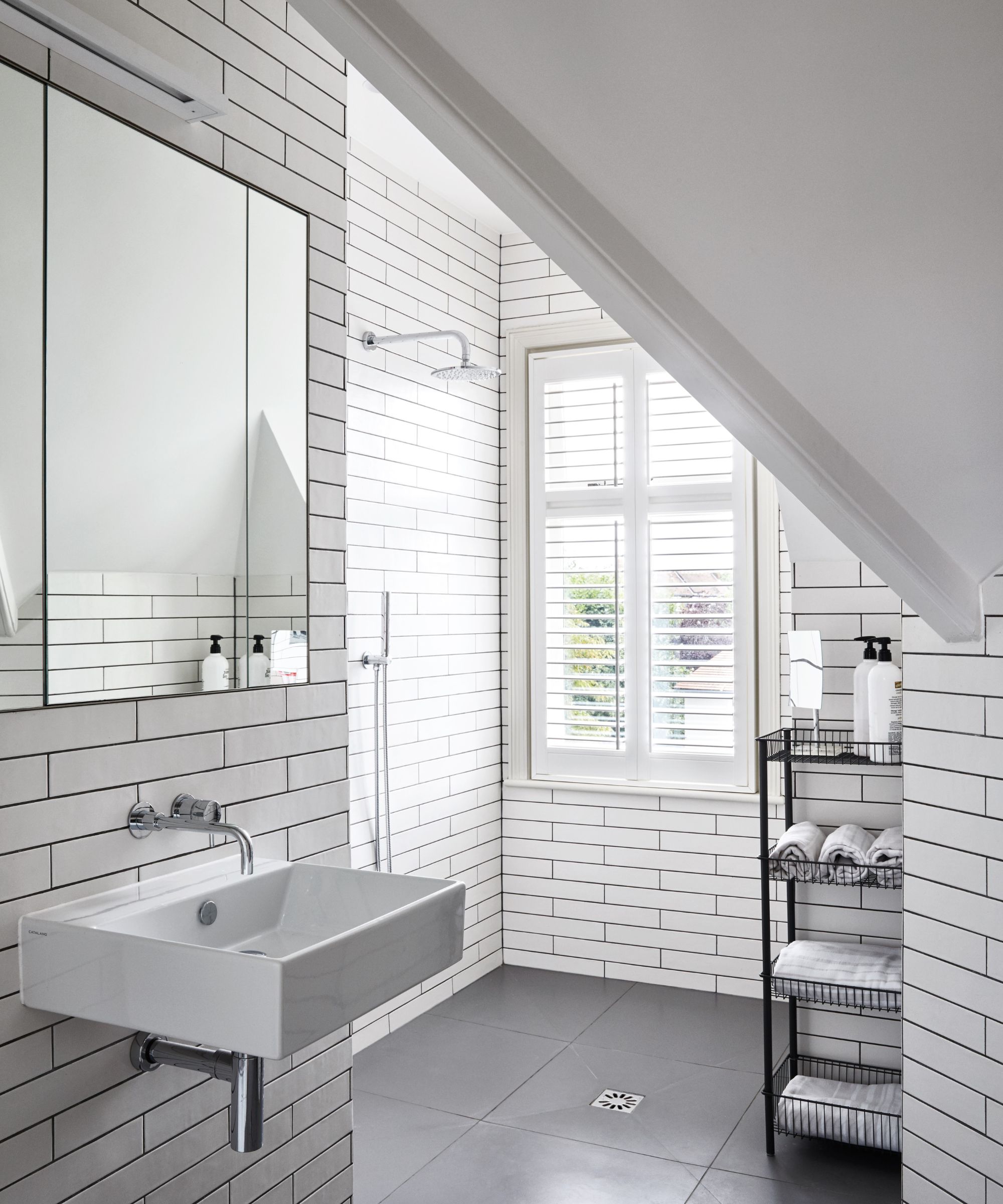 A bright white tiled attic bathroom with a white sink below a large mirror, shower, black shower caddy and large windows with white shutters.