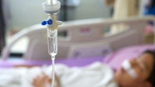 photo shows a saline drip in the foreground and a blurred out image of a child in a hospital bed in the background