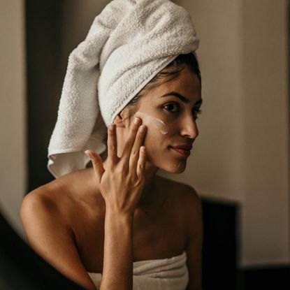 night-time skincare routine - A woman looking into the mirror with a towel on her head applying cream to her face 