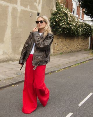 Lucy Williams wearing red wide-leg pants, an oversized distressed brown leather jacket, and a white tee, and brown sunglasses.