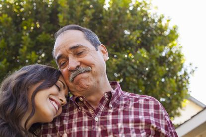 An older father smiles as his adult daughter leans her head on his shoulder. Both have their eyes closed.