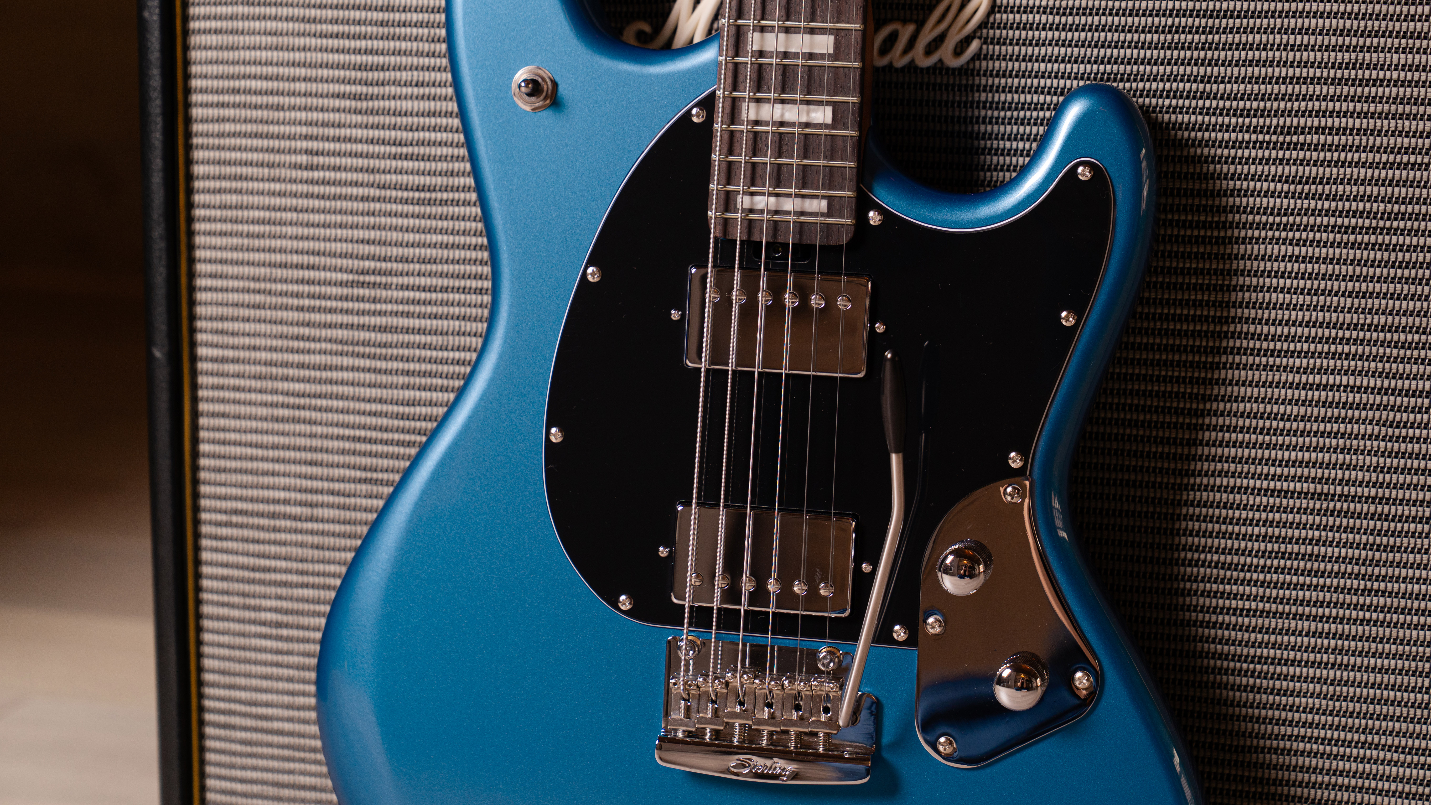 A lifestyle press image of the Sterling By Music Man StingRay Baritone SR50 in Toluca Lake Blue leaning against a Marshall amp in an empty room with concrete walls