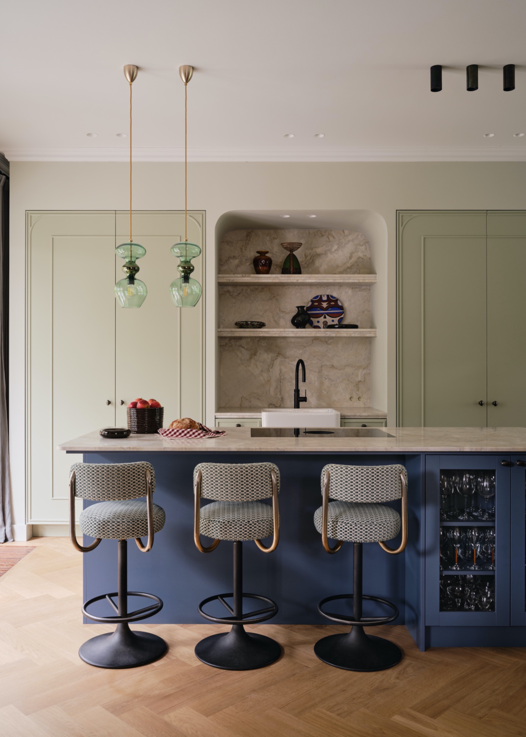 pale green cabinetry and deep blue island with green glass pendant lights, stone niche and patterned bar stools.