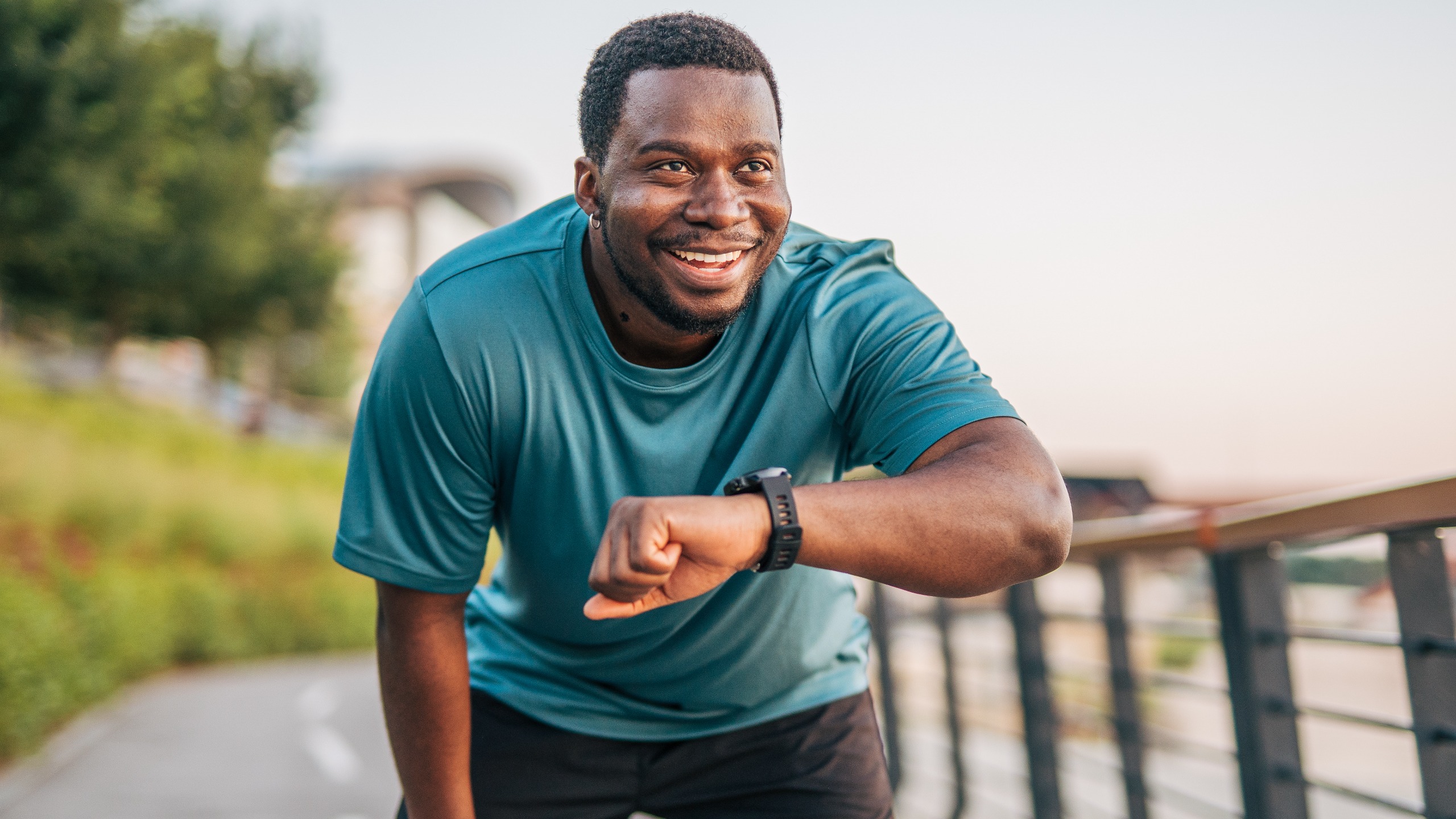 A man in running attire pauses in a bent over position, raising his left hand in front of him to look at his running watch. 