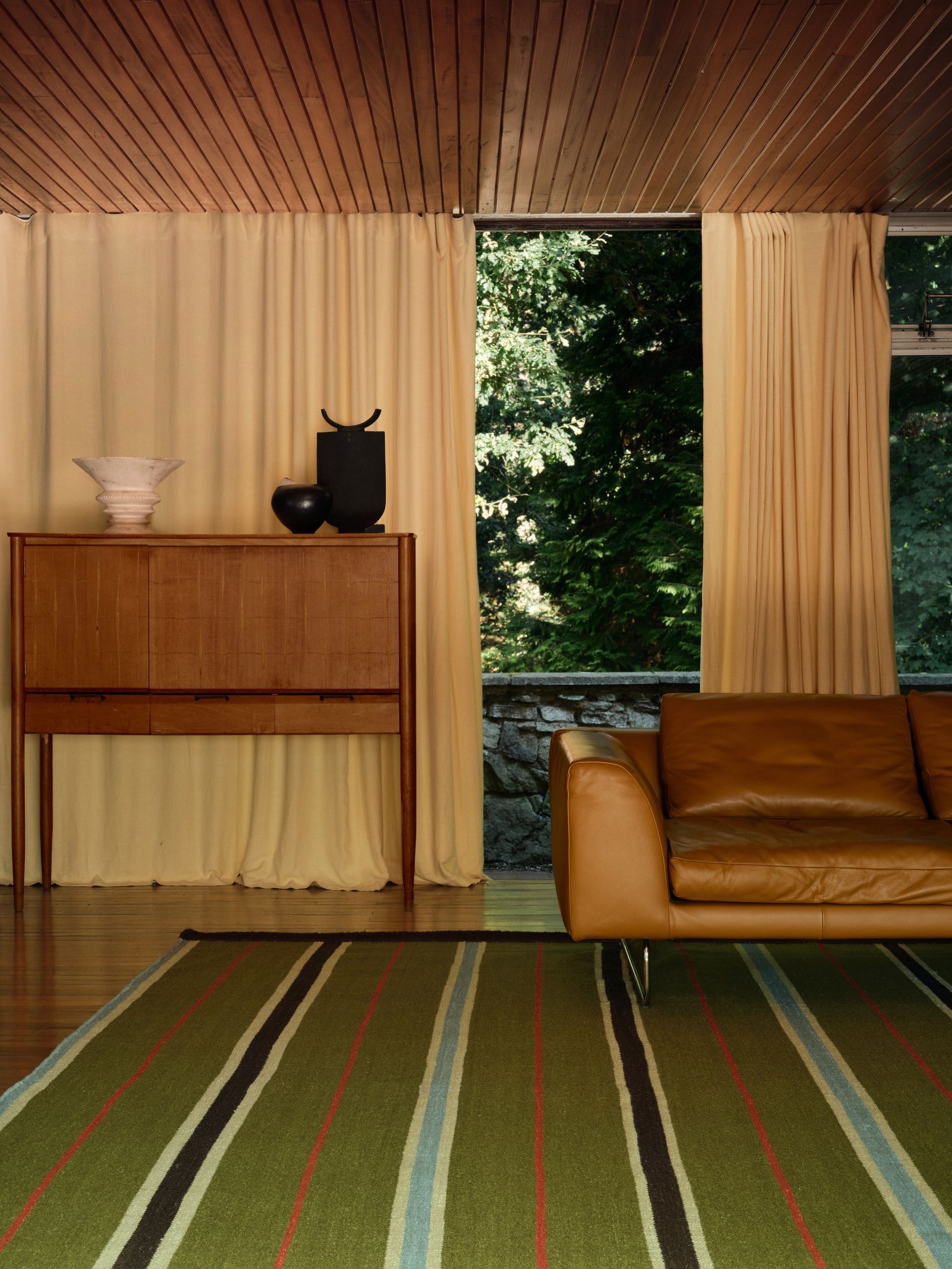 A mid-century living room where the back wall is windows with cream curtains. There is a brown leather sofa, a wooden console, and a green, red, blue, and brown striped rug.