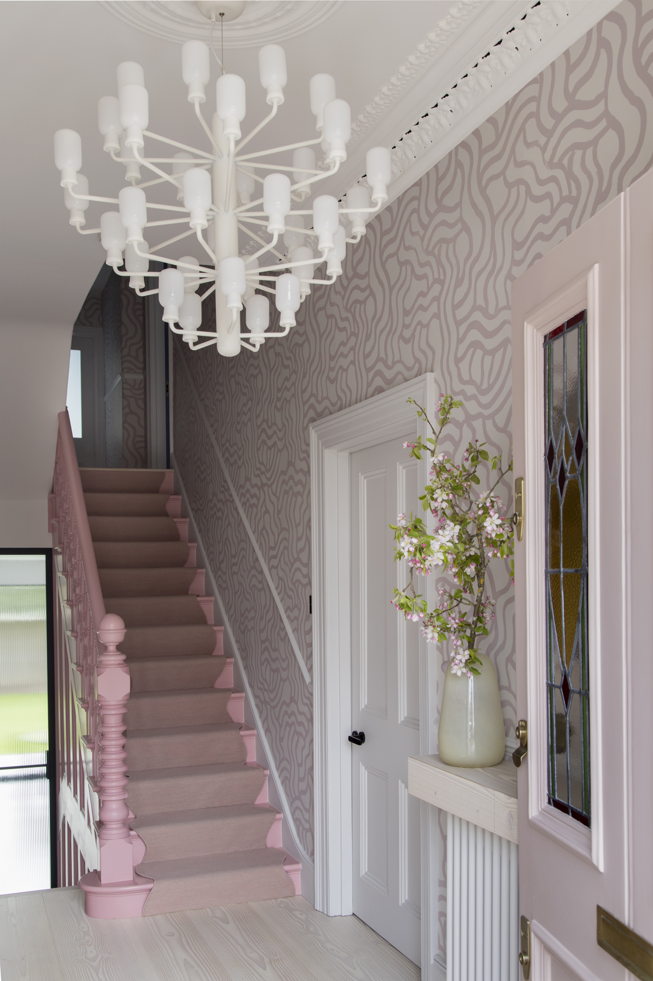 pink entryway with pink painted staircase, pink squiggly wallpaper, cornice, modern chandelier, light floorboards, a console table with a vase of pink flowers, and a pink front door