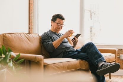 An older man sits on a couch holding an orange prescription drug bottle and a smartphone.