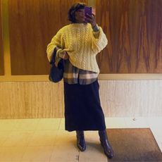 @frannfyne wears a cream cable knit jumper over a tartan shirt with a black skirt, brown knee-high boots and a black bag