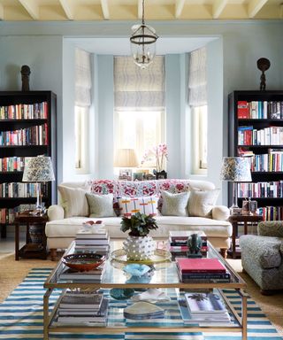 Living room with bay window, pale blue walls, built in bookcases, cream sofa with multi-coloured cushions and large square coffee table.