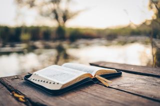 An open Bible on a wooden dock by a body of water.