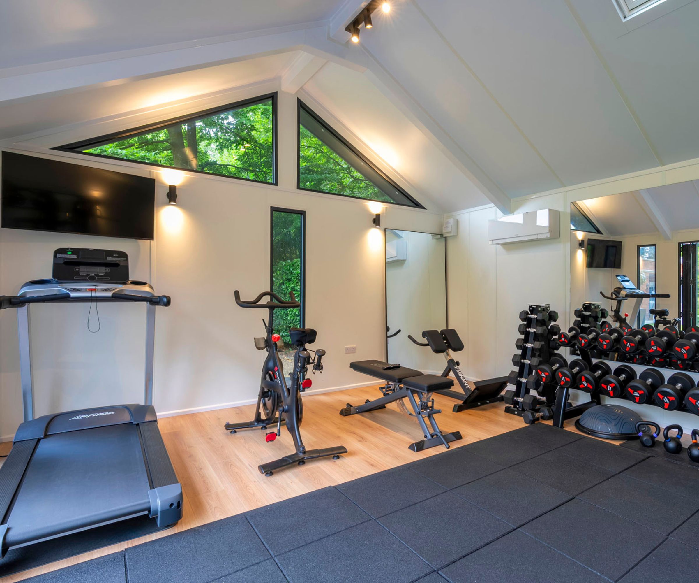 Home gym with treadmill, spin bike, weight bench and dumbbell rack under vaulted ceiling with skylights.