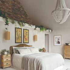 A white bedroom with high ceilings with a chandelier, an exposed brick wall and a rattan headboard 