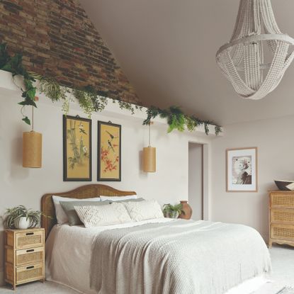 A white bedroom with high ceilings with a chandelier, an exposed brick wall and a rattan headboard