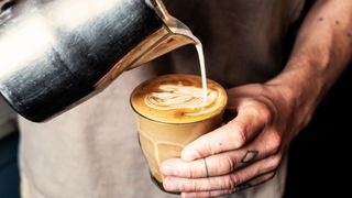Close up of person with tattooed finger pouring milk from metal jug into glass of cafe latte