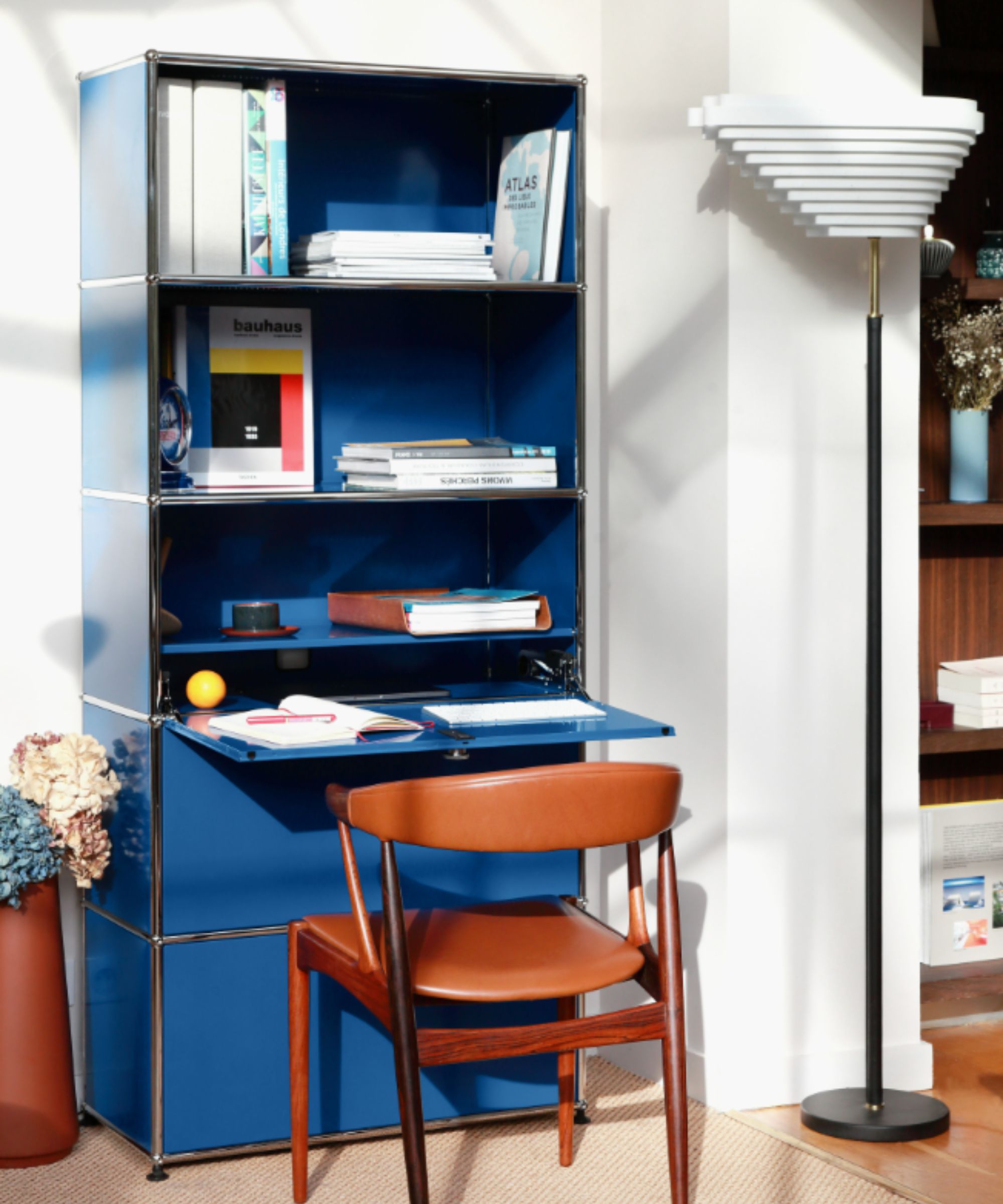 Compact home office with a bold blue shelving unit doubling as a desk, tan leather and wood chair, and sculptural floor lamp in a sunlit modern interior.