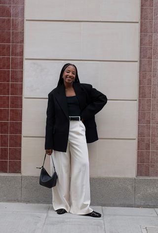 a photo showing what to wear to work in the summer with a woman wearing a black blazer over a black tank top with white trousers and flat black sandals and a black bag