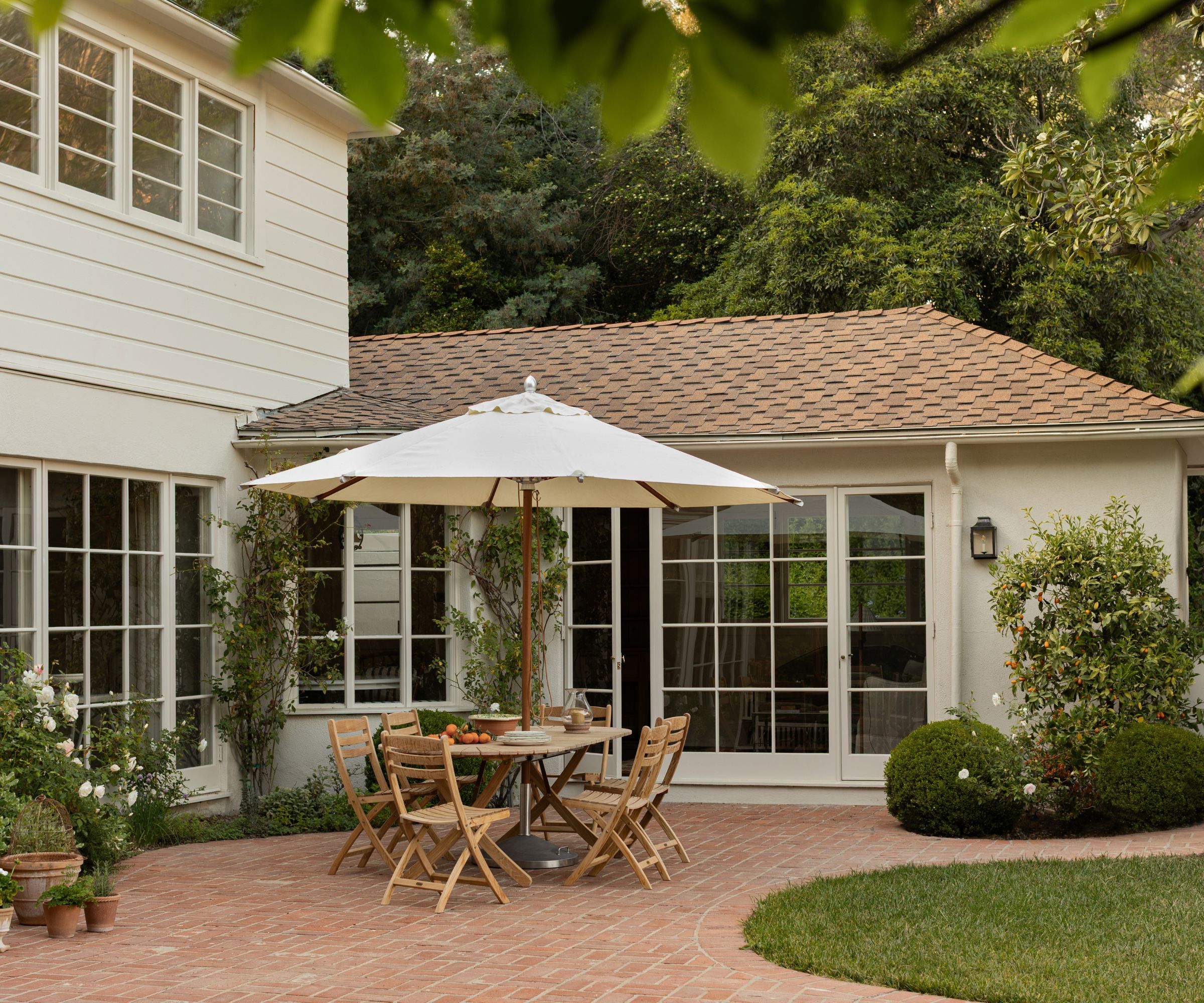 A home exterior painted warm white with a red brick roof, a patio area with a wooden table and chairs with a cream parasol, and planted shrubs.