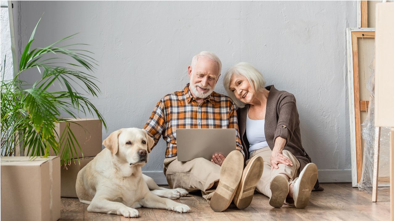 A couple considering applying for a mortgage in retirement sit on the floor of their new house with their dog and moving boxes all around them. 