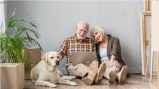 A retired couple considering a mortgage in retirement sit on the floor of their new house with their dog and moving boxes all around them. 