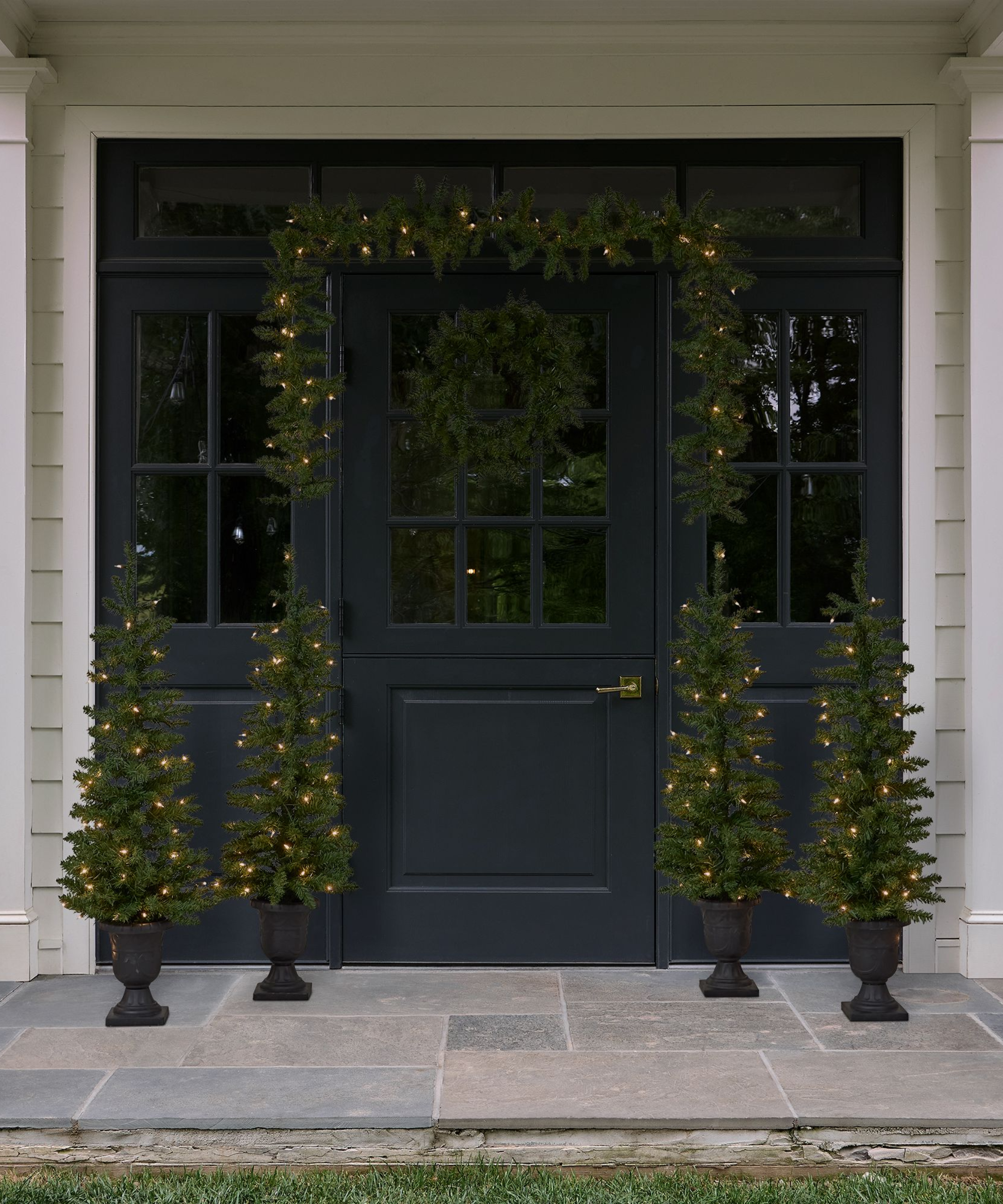 Minimalist, festive entryway featuring two pre-lit mini potted trees on either size of a navy doorway, which is decorated with a coordinating wreath and garland