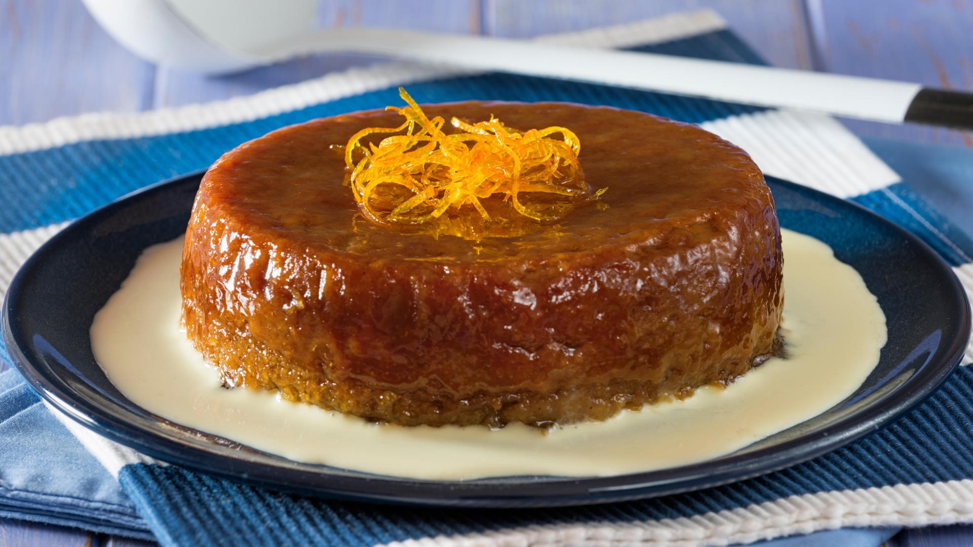 A malva pudding sits on a blue plate surrounded by a pool of custard