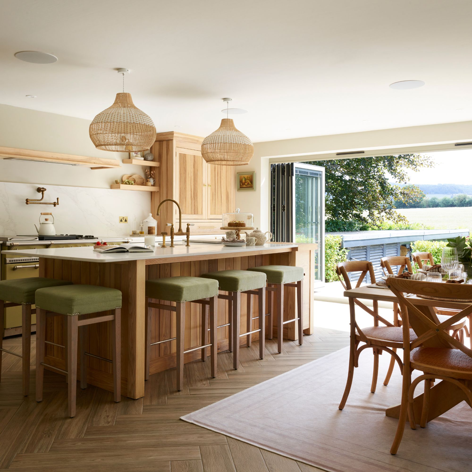 Open plan kitchen diner with wooden floors, green velvet bar stools and wooden cabinetry with marble island and wooden dining table and chairs on pale pink rug, bifold doors lead out to a garden