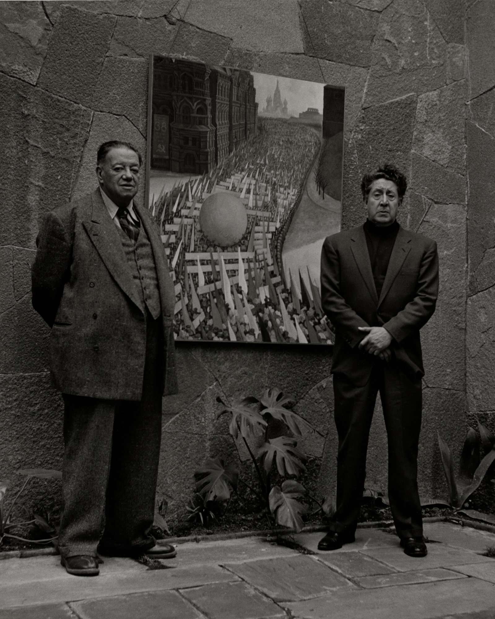 Black and white photograph of two men in suits standing either side of a framed mural depicting a crowd carrying flags through a city street.