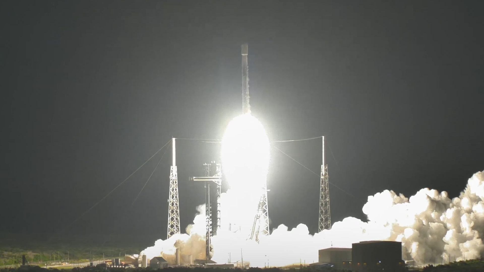 A rocket blasts into the black night sky over a launch pad, leaving a plume of white smoke in the wake of its white hot engine fire.