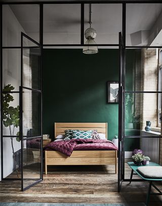 small bedroom with green focal wall, Ercol bed, black crittall doors, wooden floor, magenta quilt, turquoise cushions, pendant, industrial style space