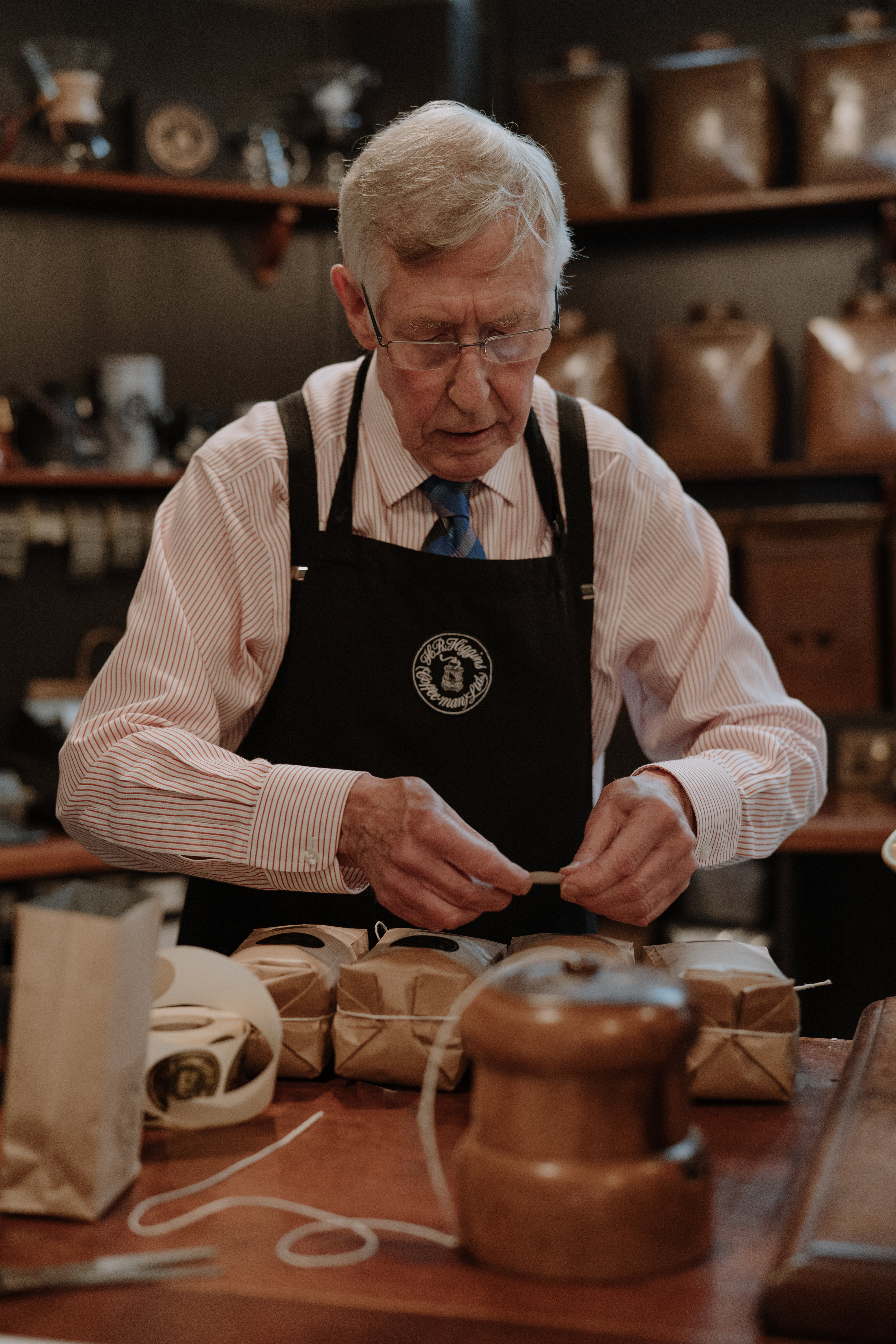 An elderly man in a black apron wrapping packets of coffee in string