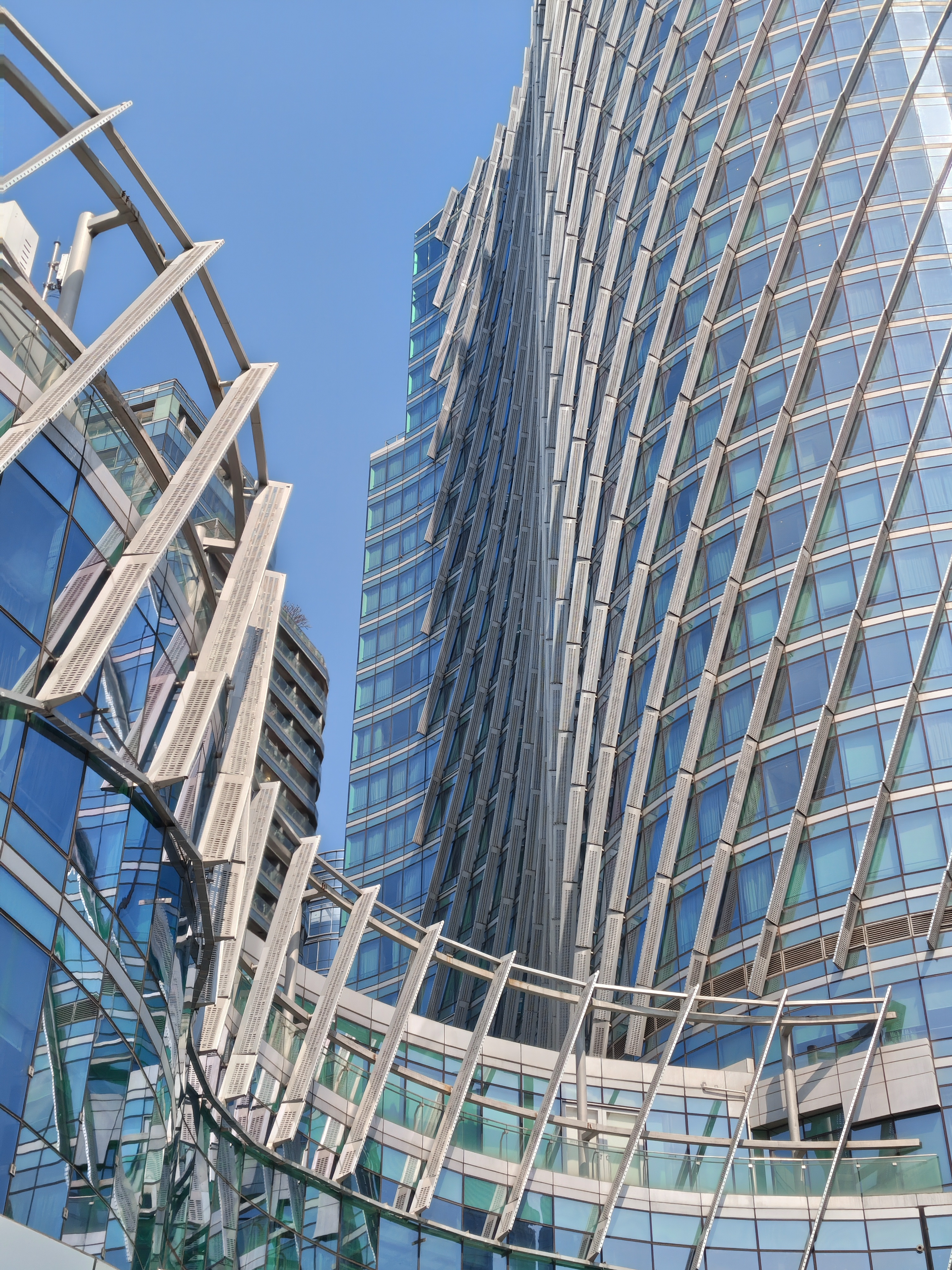 A low-angle shot of modern skyscrapers against a clear blue sky. The architecture is characterized by glass facades and prominent, diagonal metallic louvers or sunshades that create a repetitive, geometric pattern. The building on the right appears to twist as it rises, while a curved walkway structure with similar metal ribbing connects different sections in the foreground.