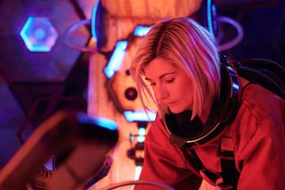 The Doctor (Jodie Whittaker) is in the Tardis, wearing an orange spacesuit with the helmet removed. She is sitting down with her head lowered, looking concerned