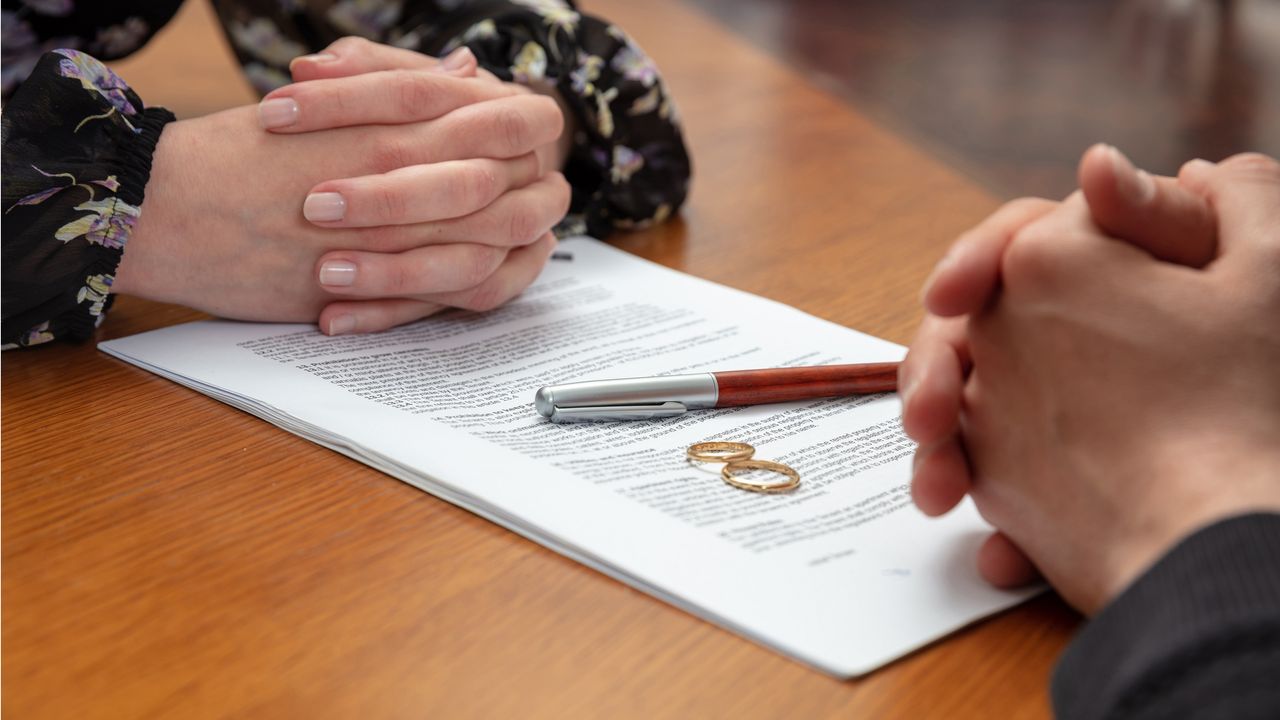 Divorce papers, wedding rings and a pen sit on a desk between the folded hands of two spouses.