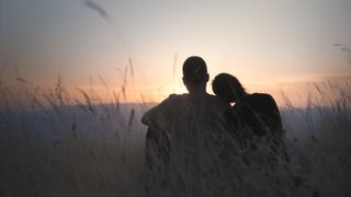 A couple in a field sit and watch the sunset.