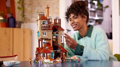 Lego The Burrow set and minifigures on a table, with a woman holding a mini owl in the background