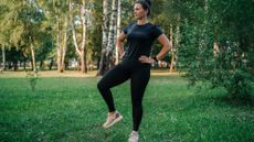 woman wearing black tshirt and leggings doing a standing hip opener exercise on grass in a park 