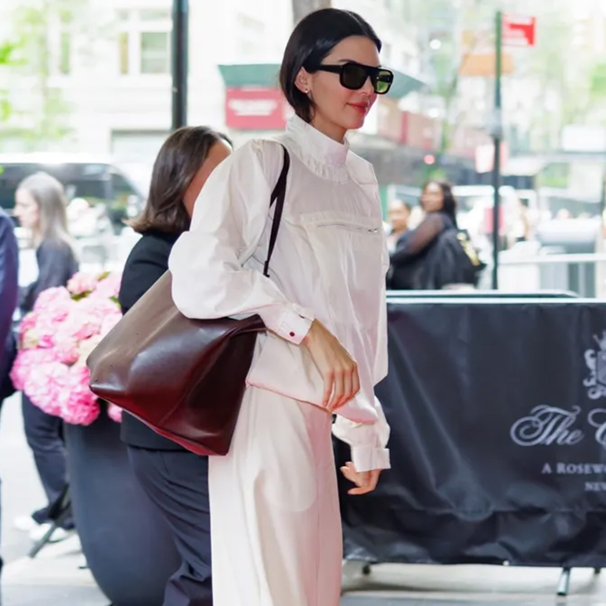 Kendall wears a white jacket, white pants, brown tote bag.