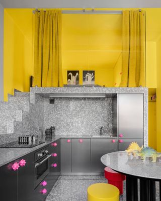 Image of a kitchen that have silver cabinetry and light gray terrazzo countertops and backsplash. The kitchen has a futuristic feel and there are stairs on the back side of the stove that lead to a yellow loft. The cabinet knows are neon pink spiky balls.