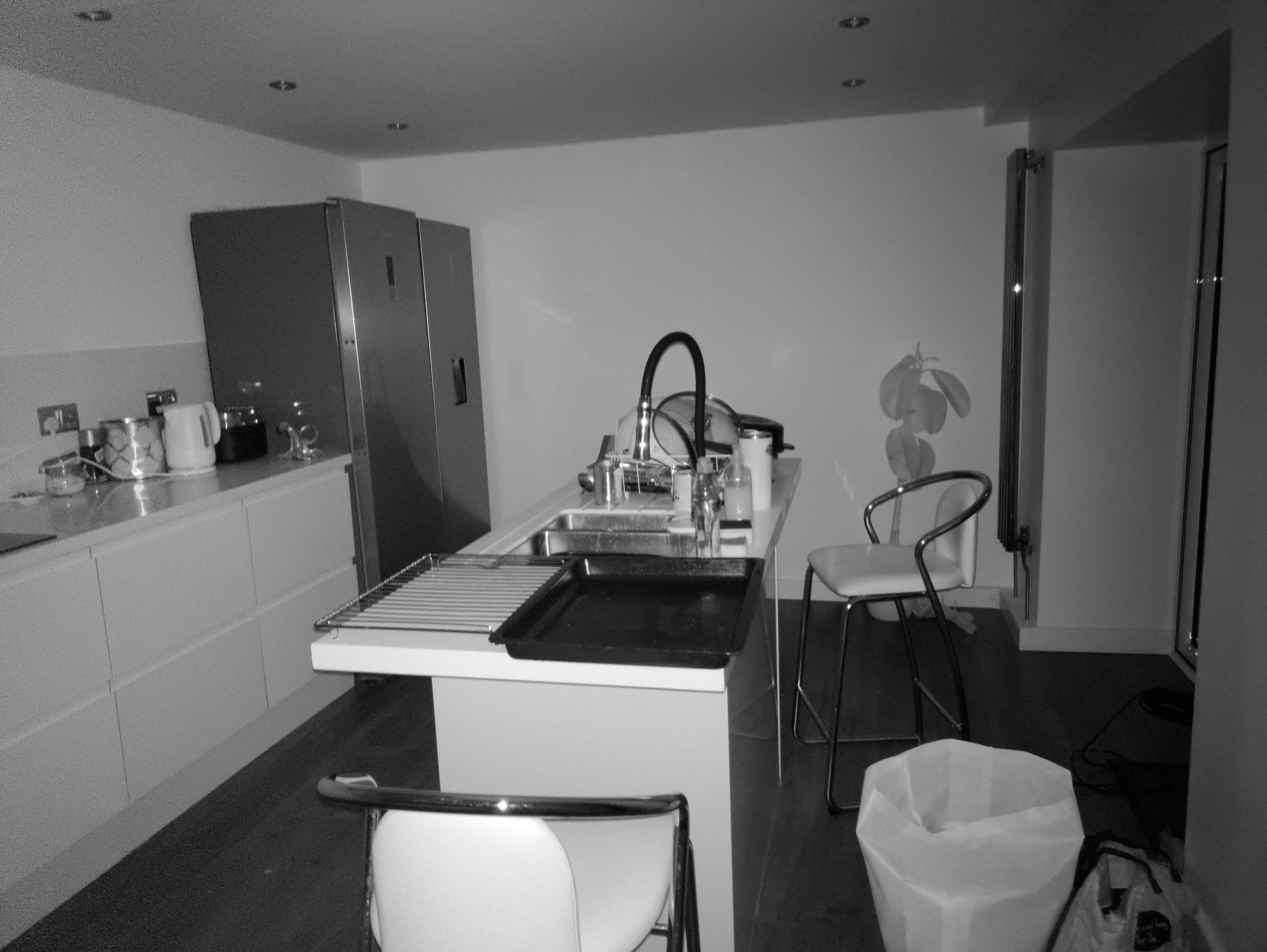 A kitchen scene featuring a modern white island with an integrated sink and faucet, two bar stools, a stainless steel refrigerator unit, and white cabinetry.