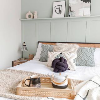 Soothing mint wall and painted paneling behind bed styled with coordinating cushions, comfy layers, and wooden tea tray.
