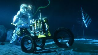 a spacesuited diver grasps a bicycle-like vehicle in an underwater facility. the diver and the vehicle are on sand. to the right is an american flag planted in the sand. an oxygen hose trails behind the diver