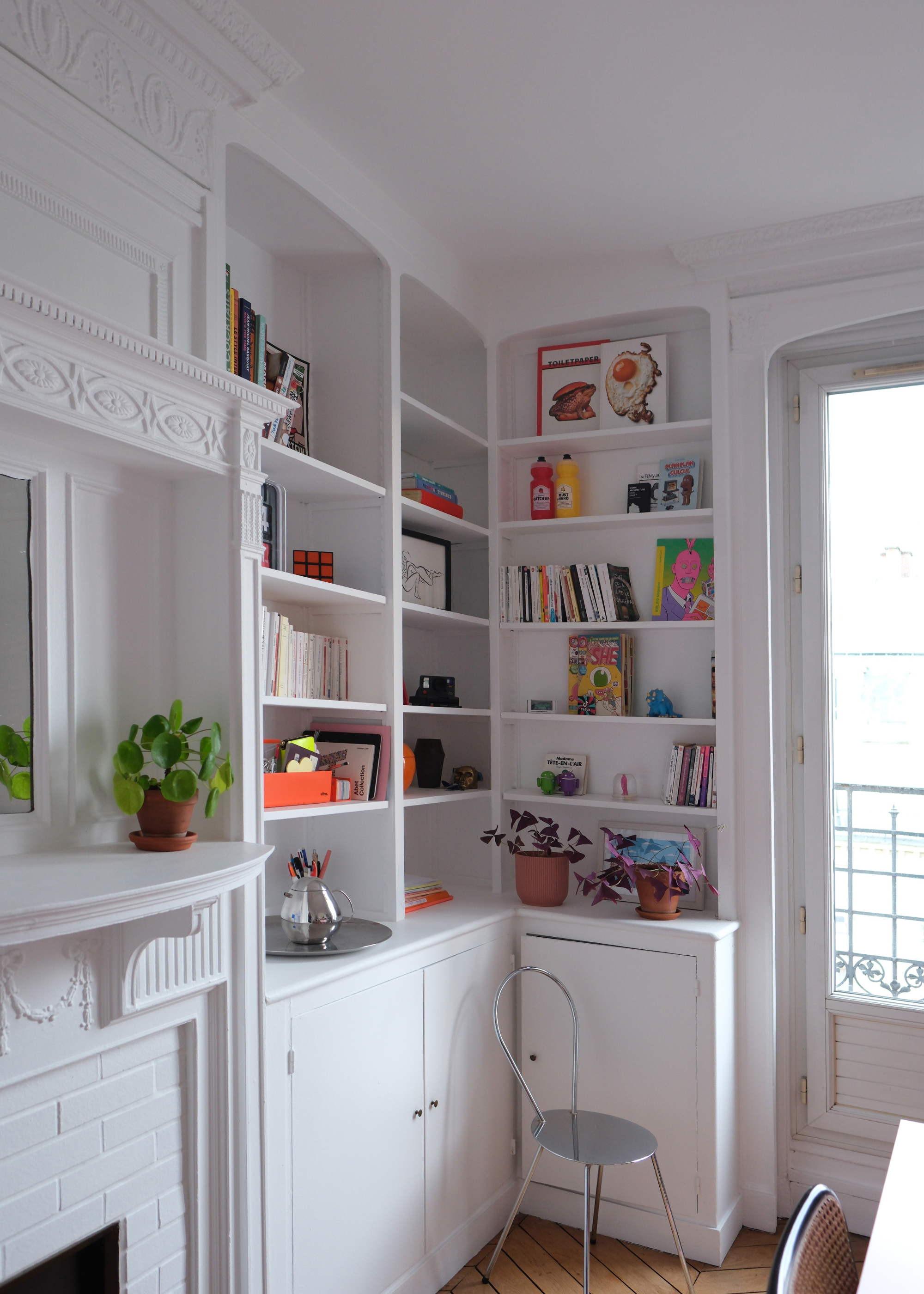 A white living room corner with shelves dotted with colorful books, prints, potted plants, and knick knacks
