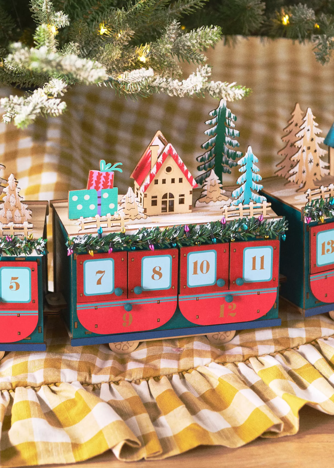 the anthropologie train advent calendar with red carriages with christmas houses, trees and presents on top of the carriages sitting on top of a gingham christmas tree skit with some frosted branches of the tree touching over the top of the train carriage