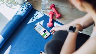 Woman using dumbbells and workout app on living room floor with yoga mat and fitness tracker watch