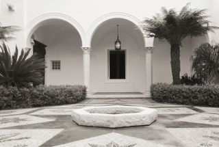 Black and white image of Von Romberg House, Montecito, California, 1938, white building with arch design and stone columns, small windows, hanging outside lantern light, palm tree, shrubs and hedges, stone centre piece in the driveway and star design stone floor