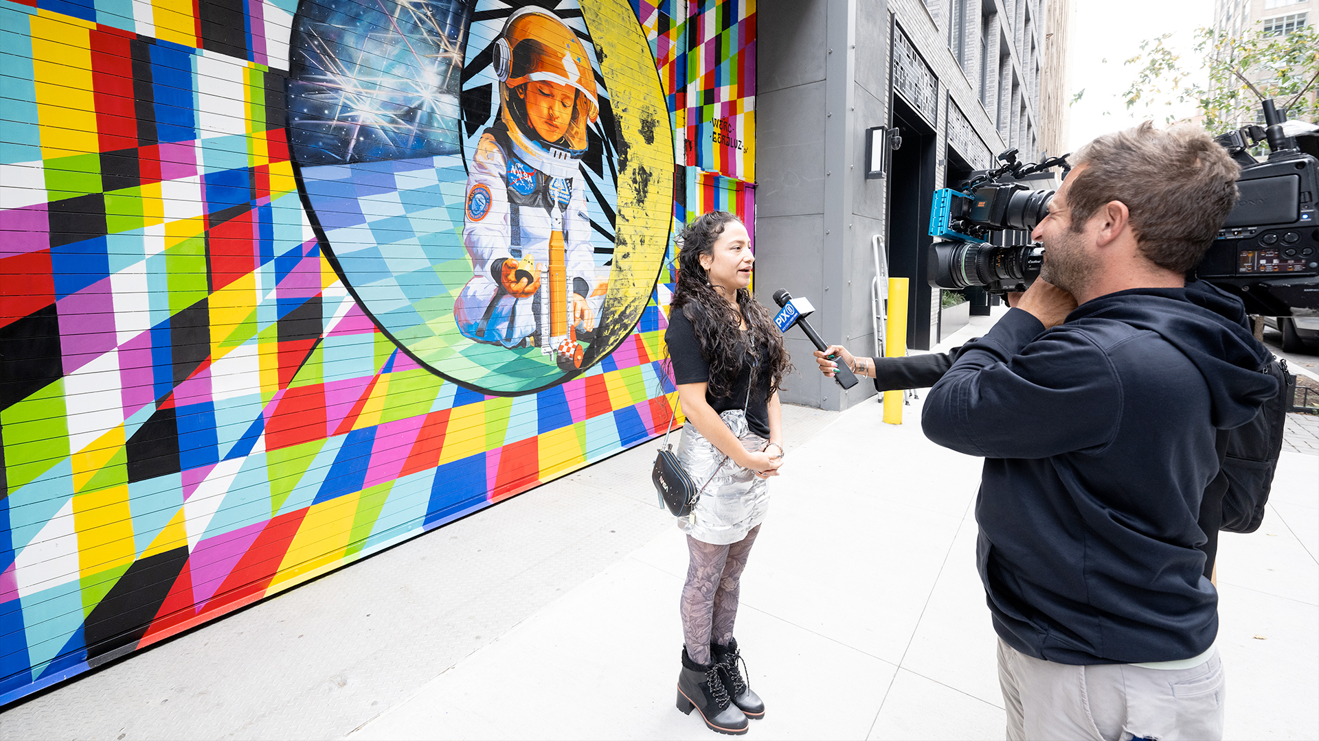 a woman is interviewed standing in front of a colorful space exploration mural