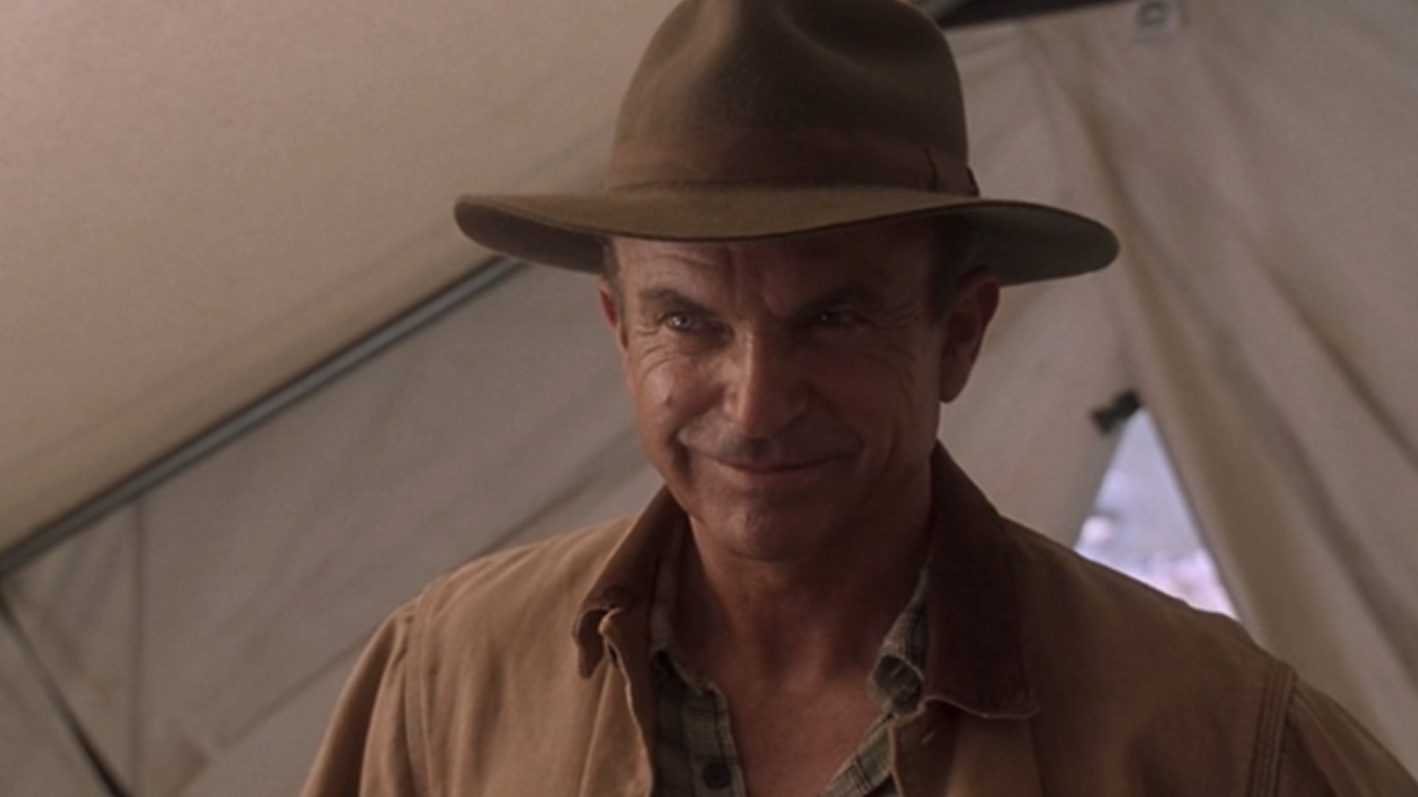 Sam Neill smiling in a tent, wearing a brown fedora in Jurassic Park III