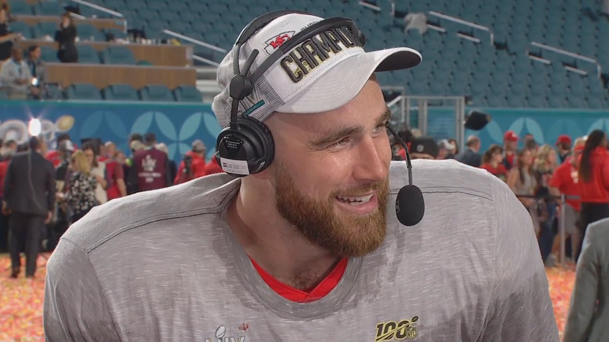 Travis Kelce is shown wearing a gray T-shirt over his football jersey and white cap, giving an interview immediately following the Chiefs' Super Bowl win on February 11, 2024.