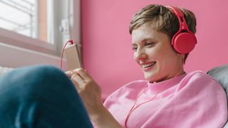 A woman wearing pink headphones smiles as she looks at her phone.