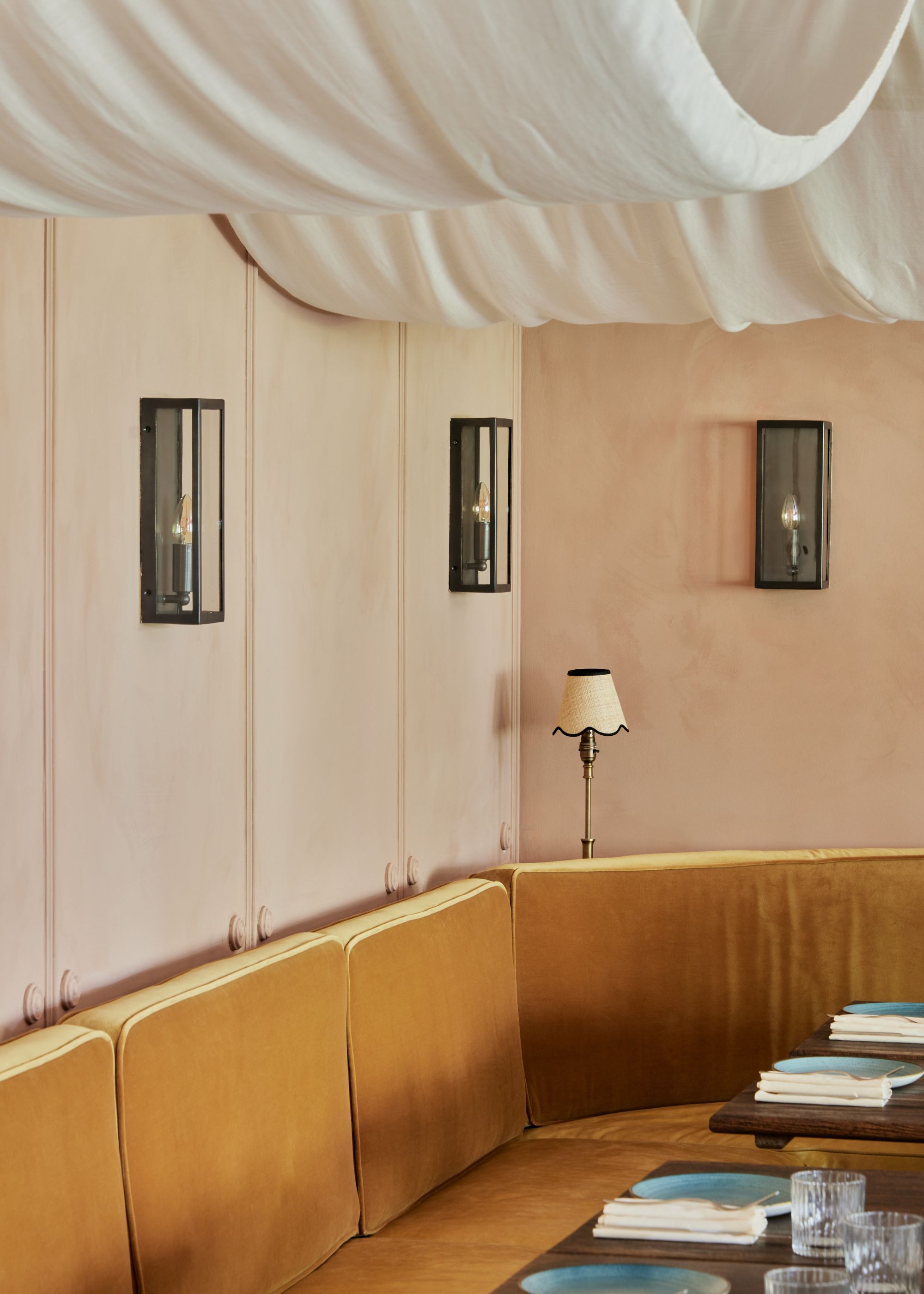 A restaurant seating area with an ochre banquette. Plaster colored walls with three black wall sconces, and white billowy draped fabric on the ceiling.